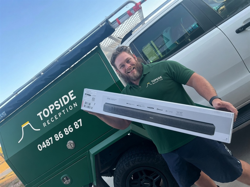 Nick from Topside Reception grinning beside the green Topside ute, holding a boxed sound bar